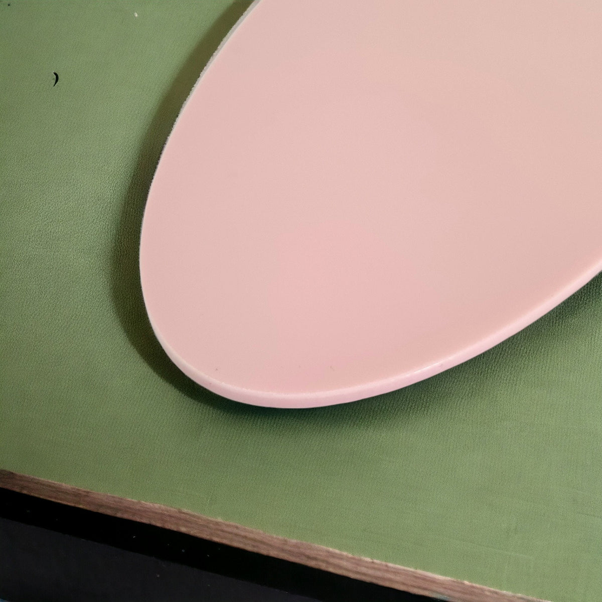 A smooth, pale pink oval acrylic worktop saver resting on a textured sage green surface.