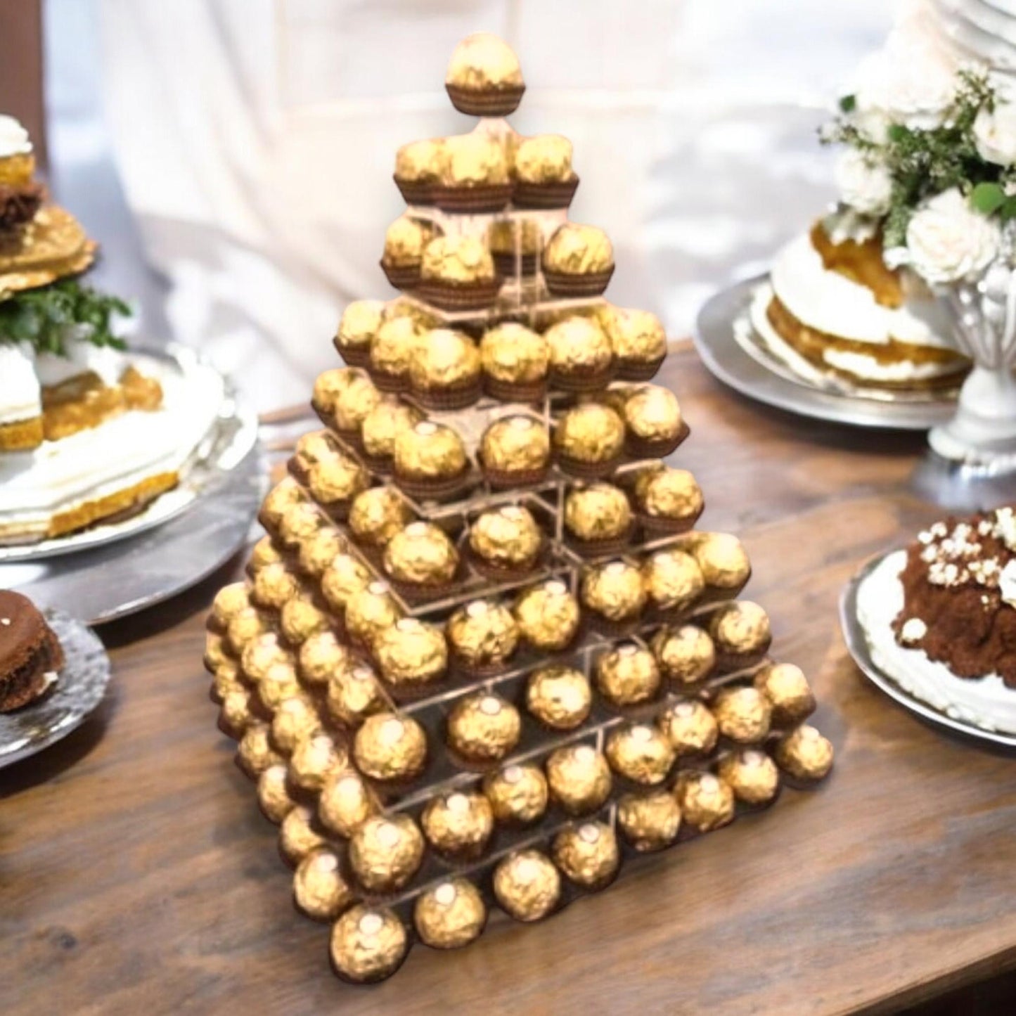 Tiered square acrylic pyramid display stand holding rows of gold-wrapped chocolates on a wooden table.