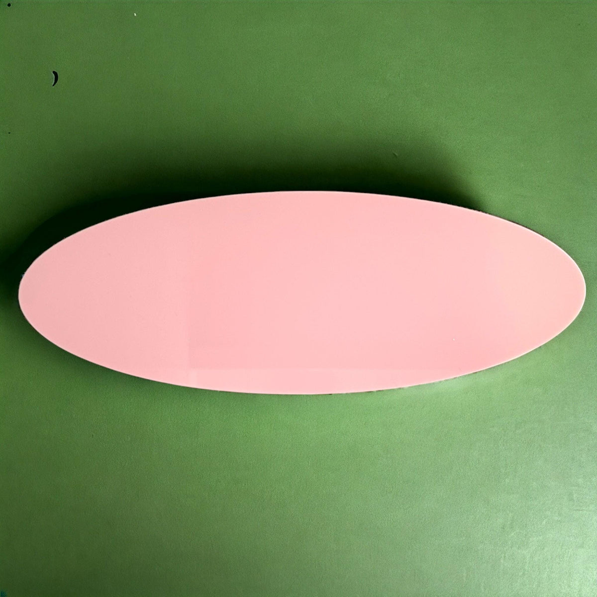 A smooth, oval-shaped light pink acrylic table runner placed on a solid green background.