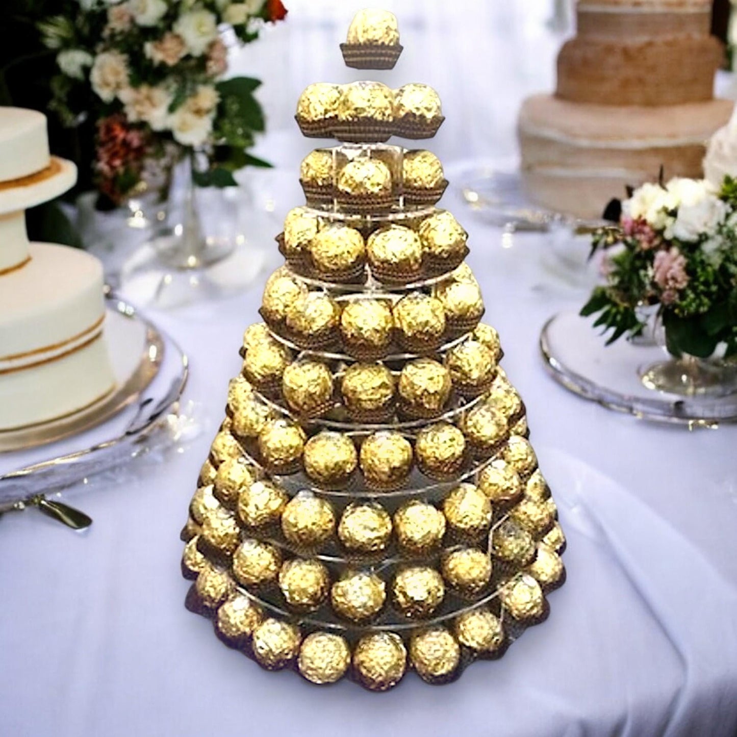 Multi-tier acrylic pyramid display stand holding golden chocolates, set on a white tablecloth at a wedding reception.