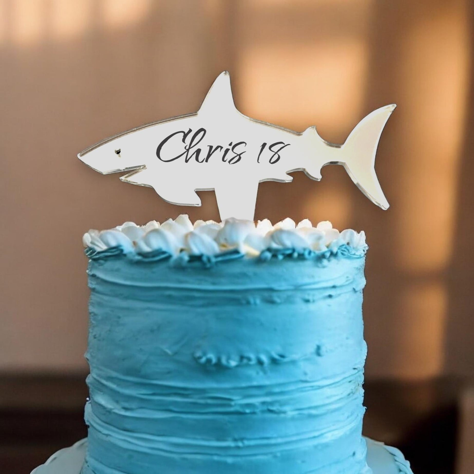 Mirrored shark cake topper engraved with "Chris 18" in black cursive, sitting on a blue frosted cake.