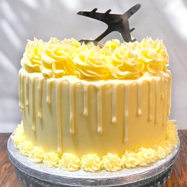 Shiny silver airplane cake topper on a yellow cake with white chocolate drip and buttercream swirls.
