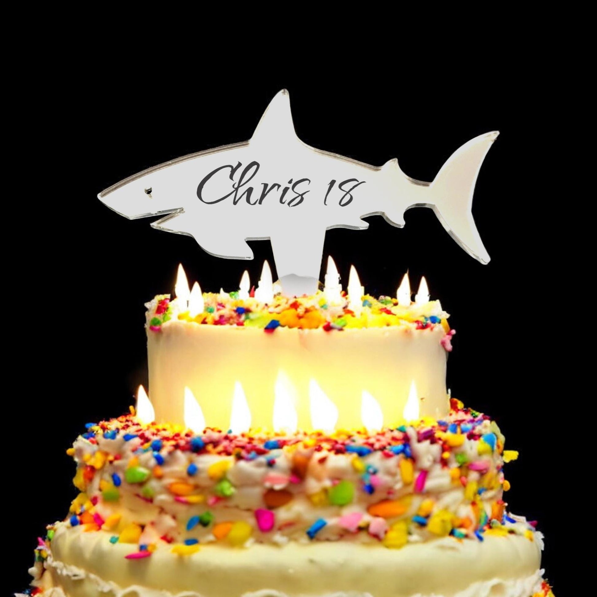 Mirrored shark-shaped acrylic cake topper engraved with "Chris 18" atop a multi-tiered sprinkle cake with candles.