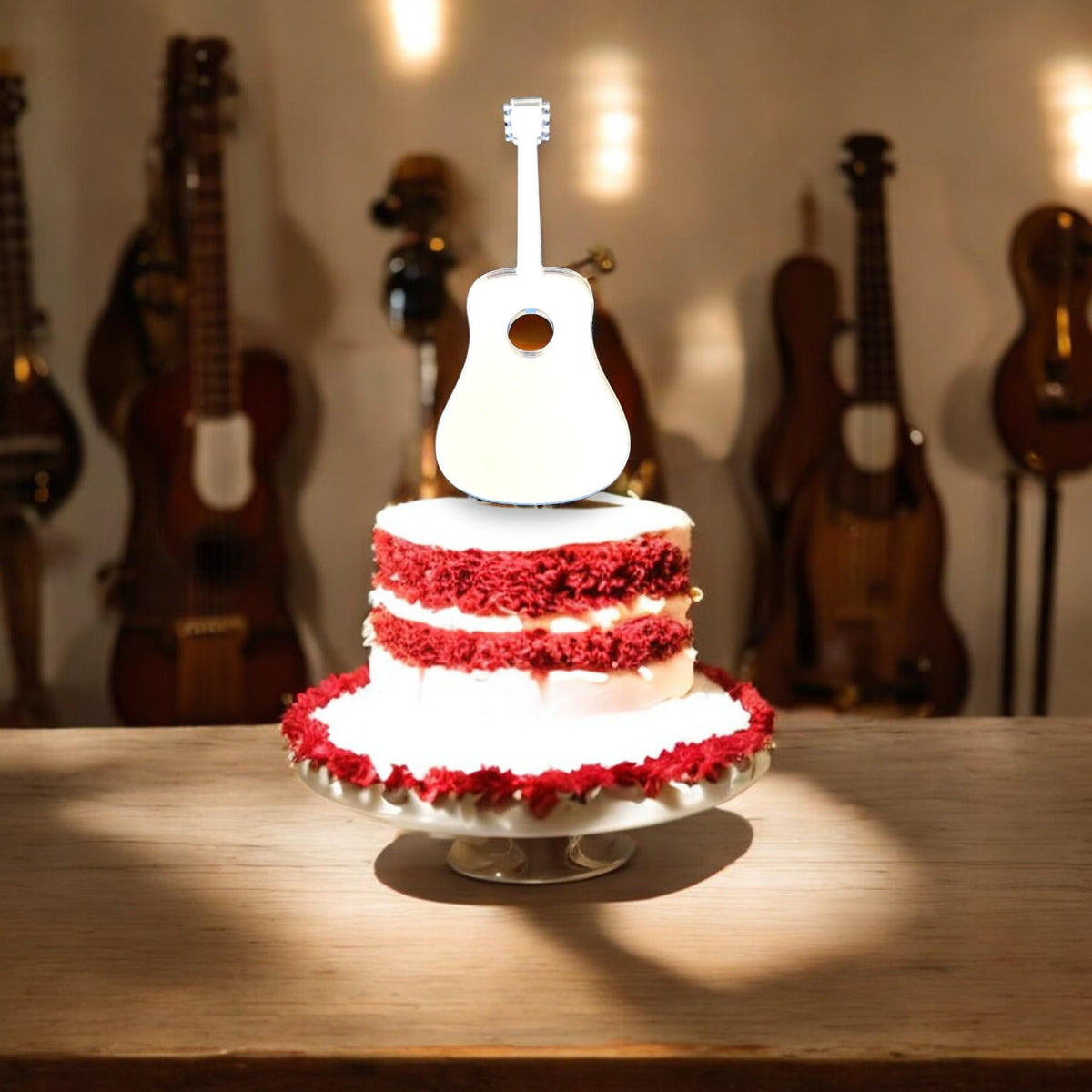 White acoustic guitar-shaped cake topper on a red velvet cake, with out-of-focus guitars in the background.