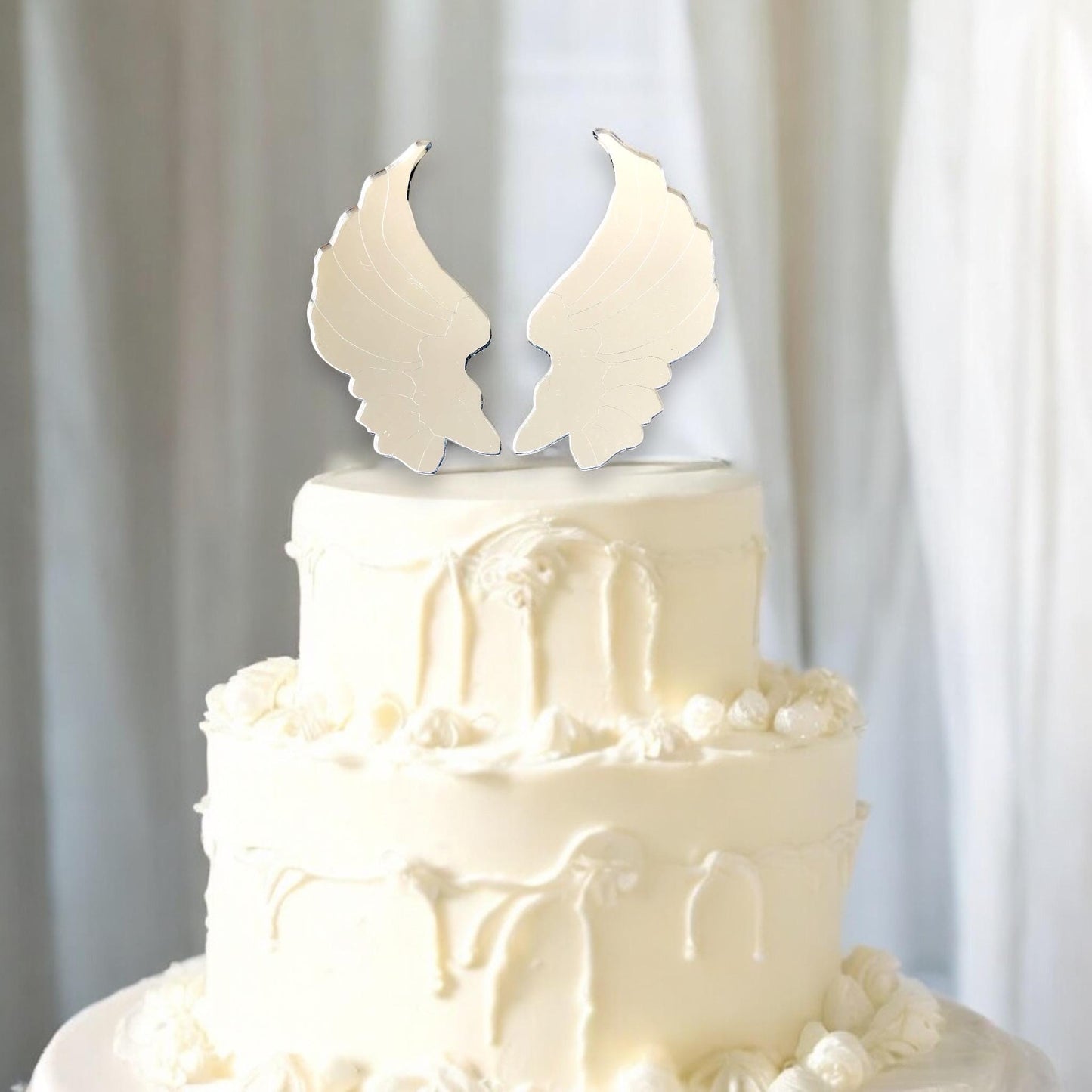 Reflective silver, mirror-finish angel wing cake topper placed on top of a two-tier white cake.