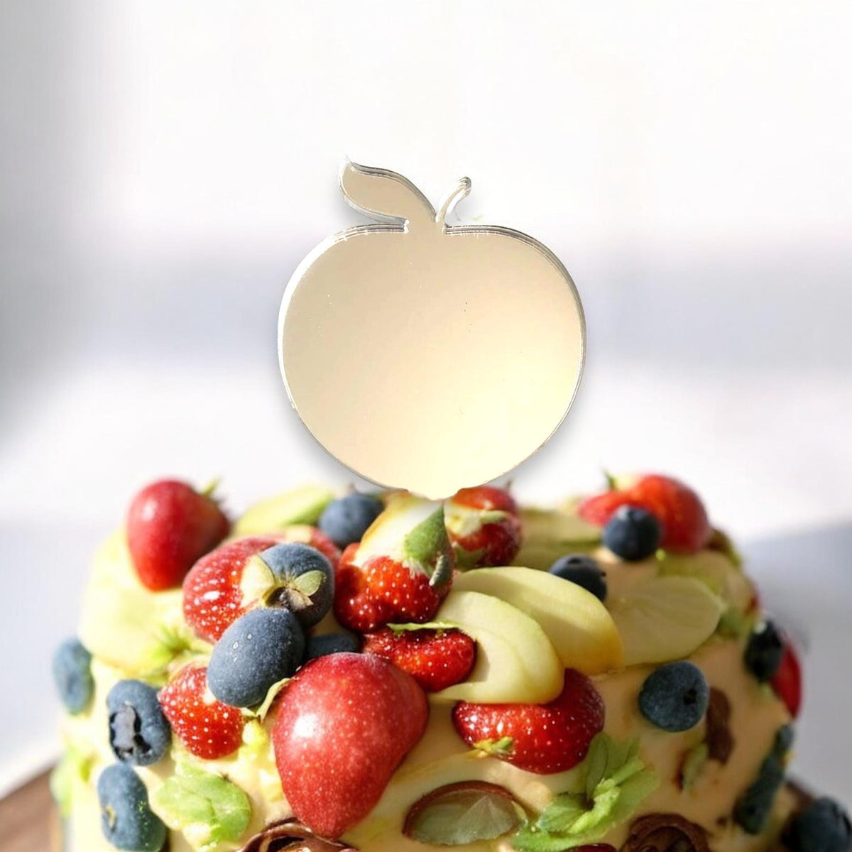 Mirror-gold acrylic apple-shaped cake topper on a fruit-covered cake.