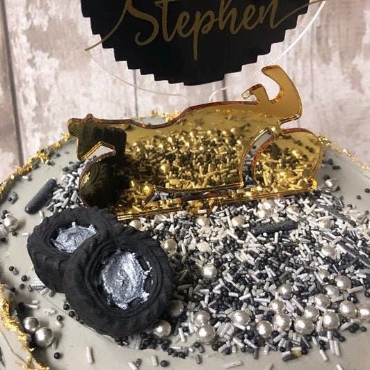 Gold mirrored acrylic motorcycle cake topper featuring the name "Stephen" on a black sign, surrounded by cake sprinkles.