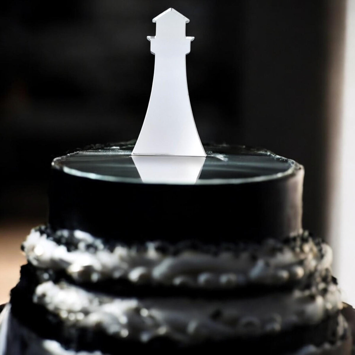 A shiny silver mirrored lighthouse cake topper on a black tiered cake with white ruffled decorations.