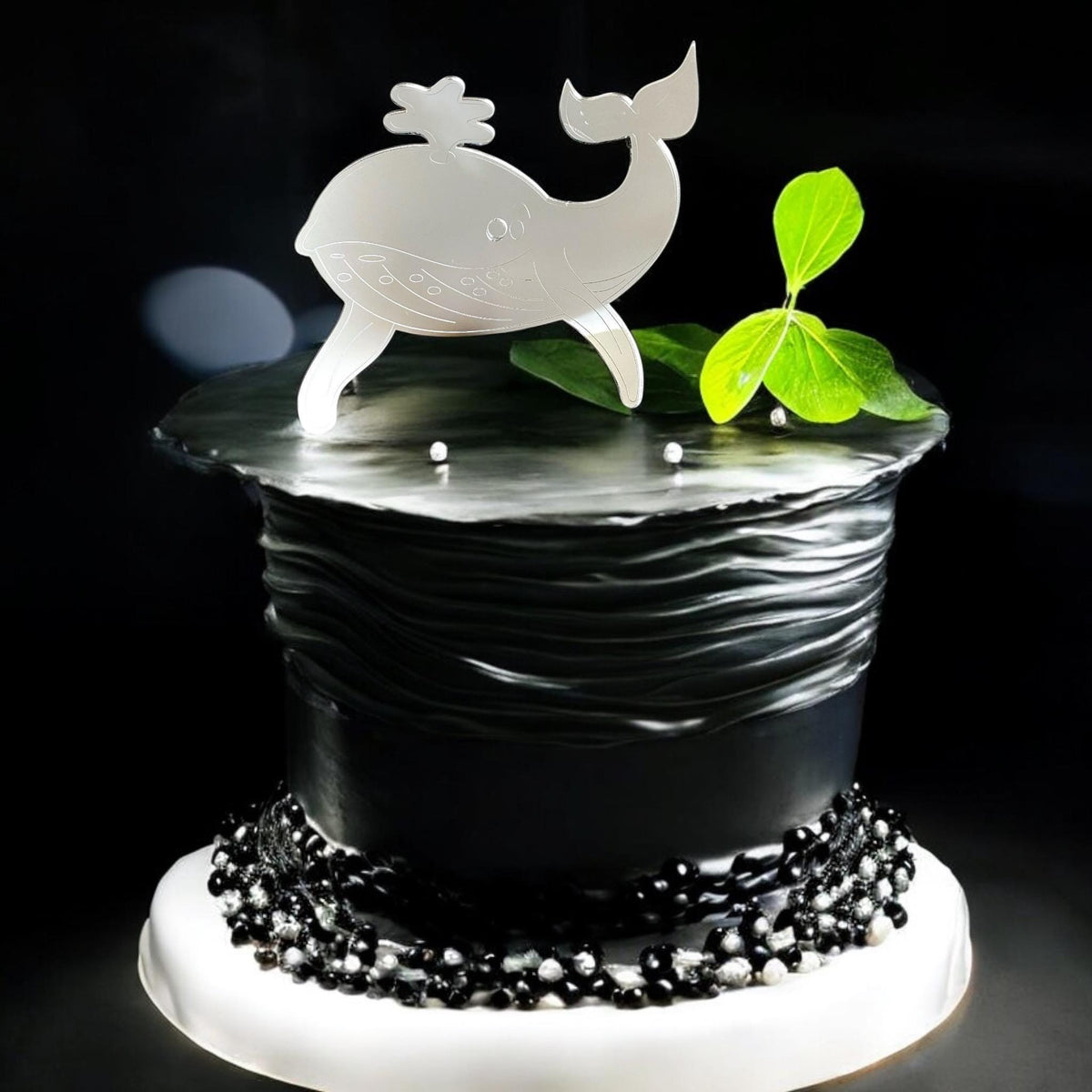 Reflective silver whale acrylic cake topper on a textured black cake decorated with pearls and green leaves.
