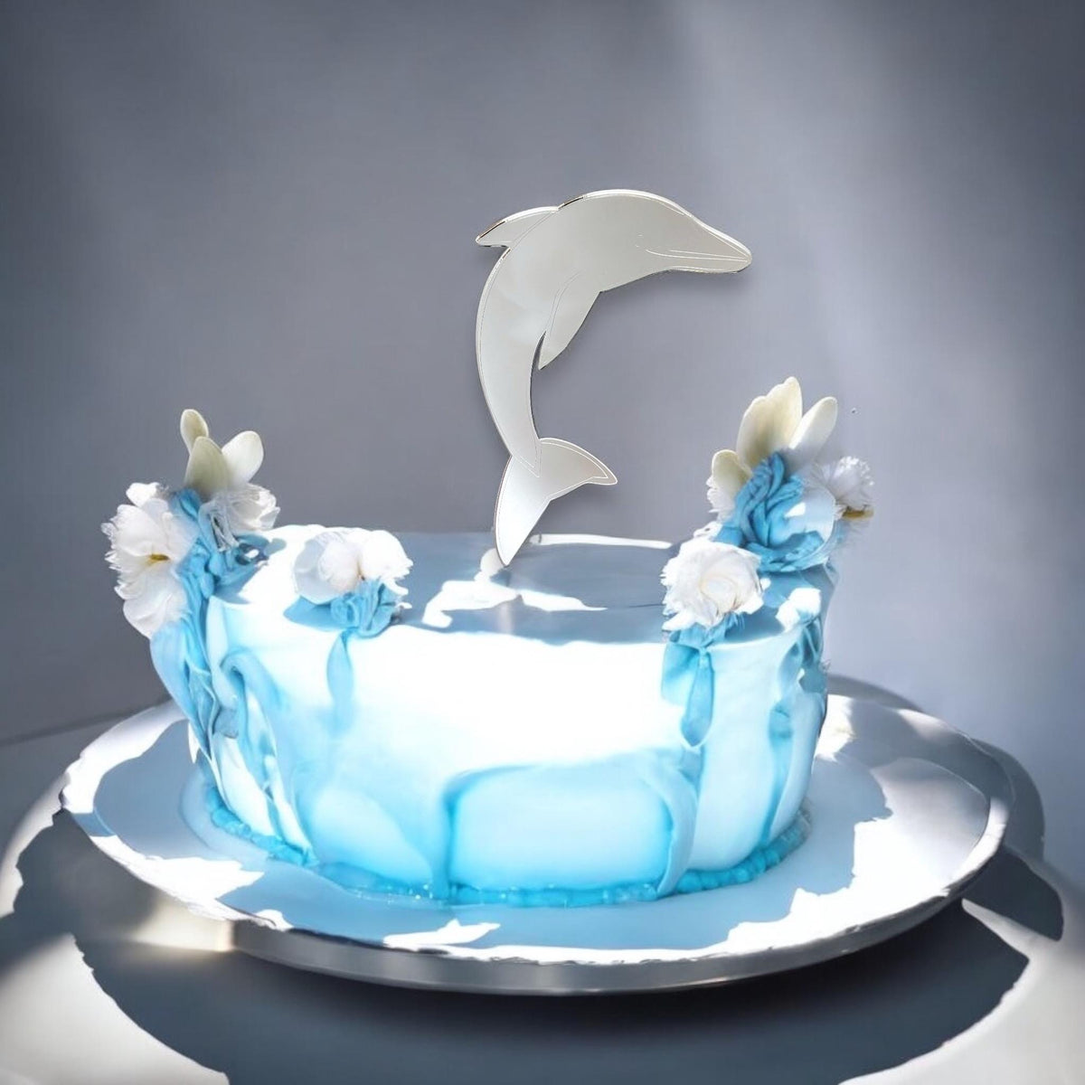 A reflective silver acrylic dolphin cake topper sits atop a light blue cake decorated with white and blue flowers.