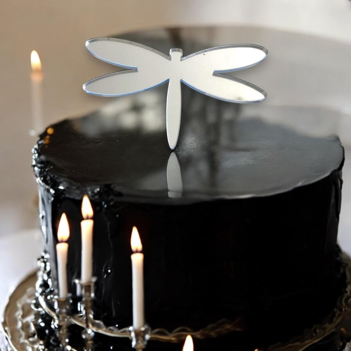 A shiny, silver, mirrored dragonfly cake topper centered on a round, glossy black cake with lit white candles.