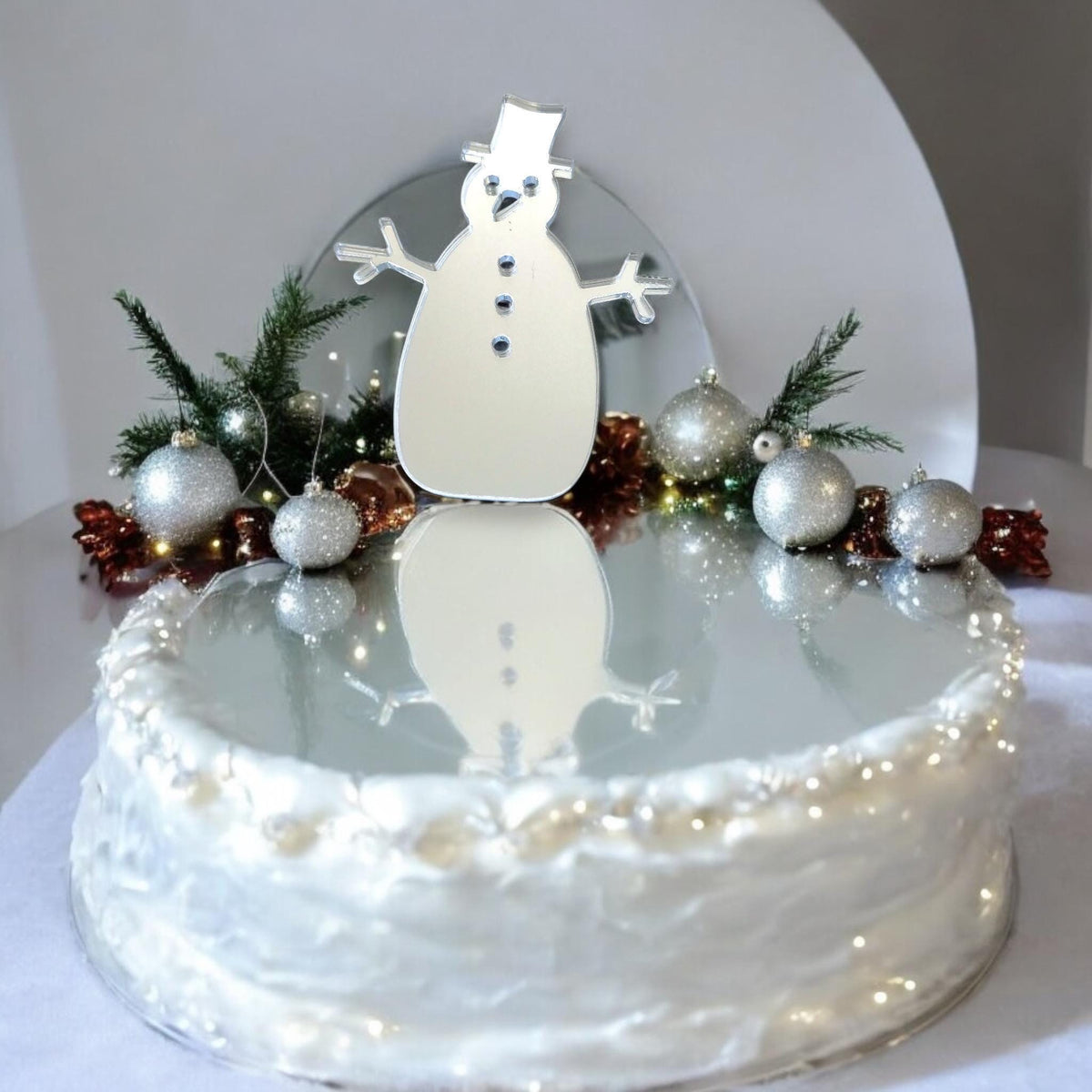 Silver mirrored snowman cake topper on a white, frosted cake decorated with silver ornaments and greenery.
