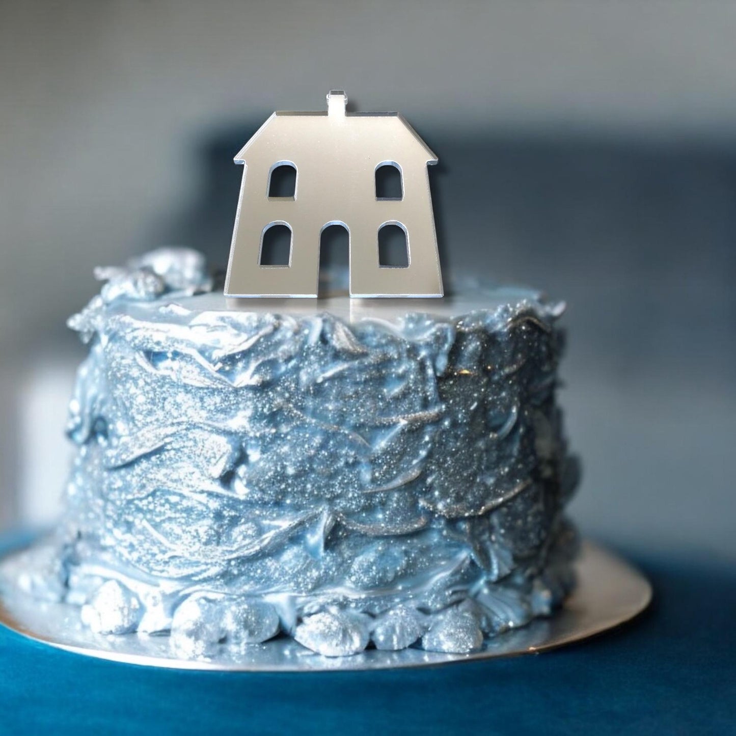 A silver mirror-finish dolls house cake topper by SuperCoolCreations on a textured, glittery blue frosted cake.