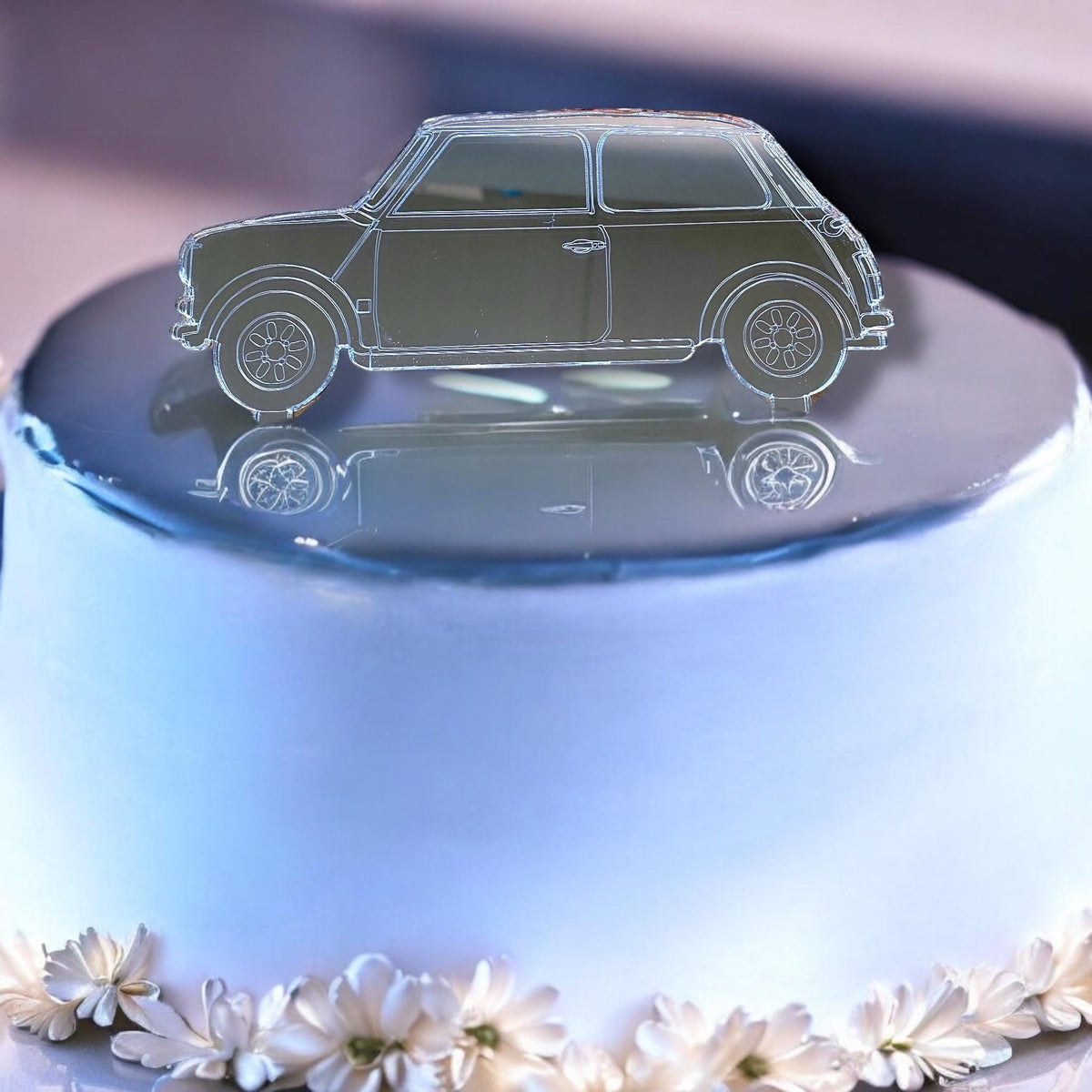 Mirror-finish silver acrylic cake topper shaped like a classic Mini car, resting on a white cake with floral trim.