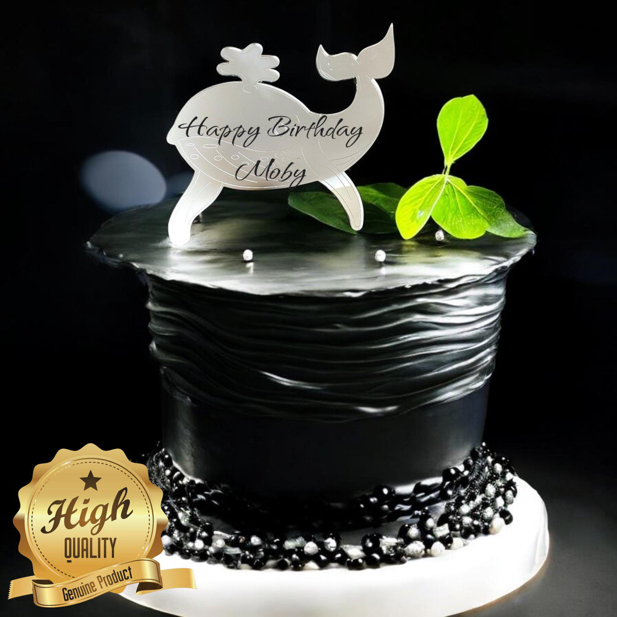 Silver mirrored acrylic whale cake topper reading "Happy Birthday Moby" on a black textured cake.