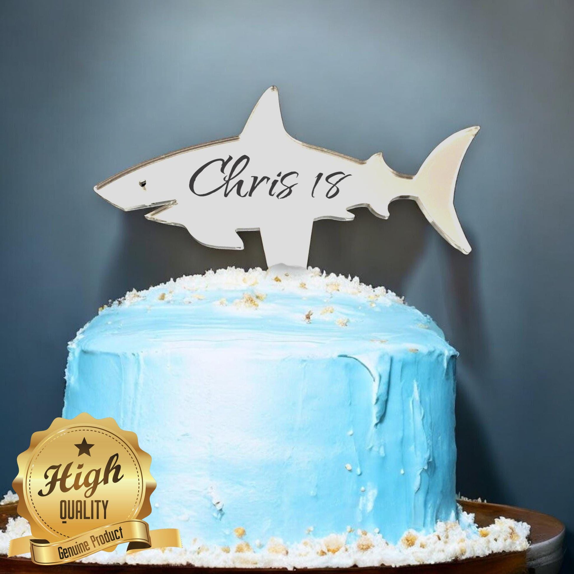 Mirror acrylic shark cake topper engraved with "Chris 18" on a blue frosted cake.