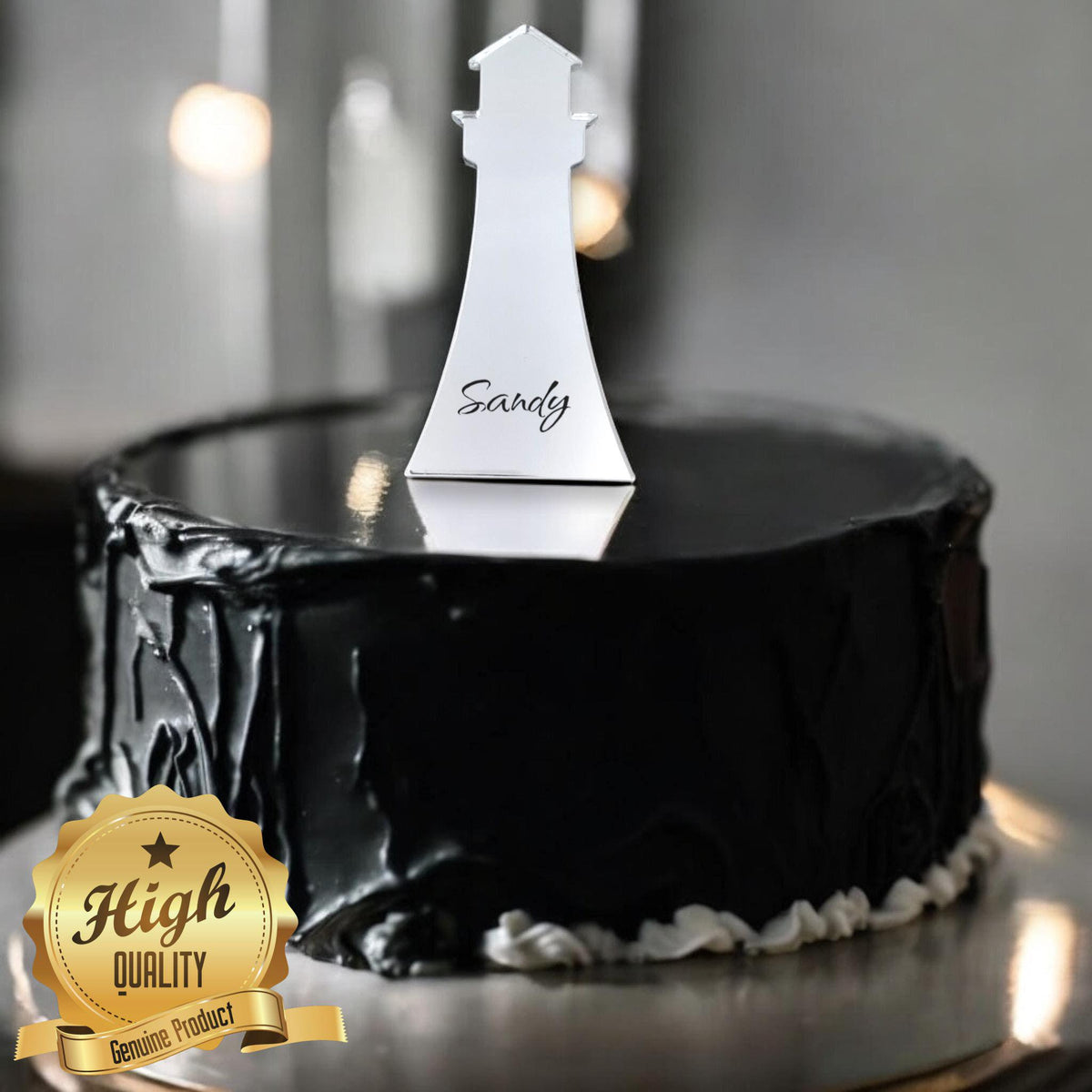 Silver lighthouse cake topper personalized with the name "Sandy," centered on a black frosted cake.