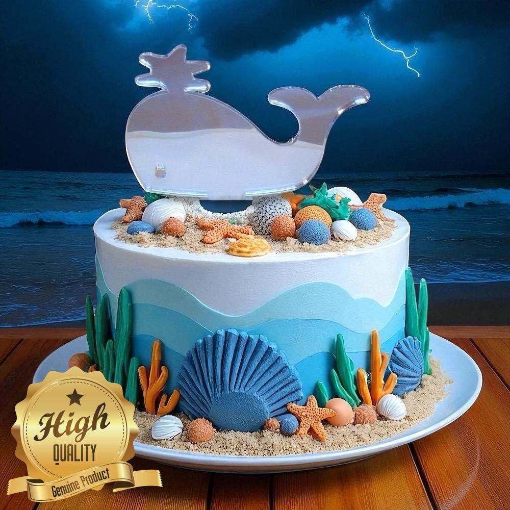 Clear acrylic whale cake topper on an ocean-themed cake decorated with fondant seashells, coral, and sand.