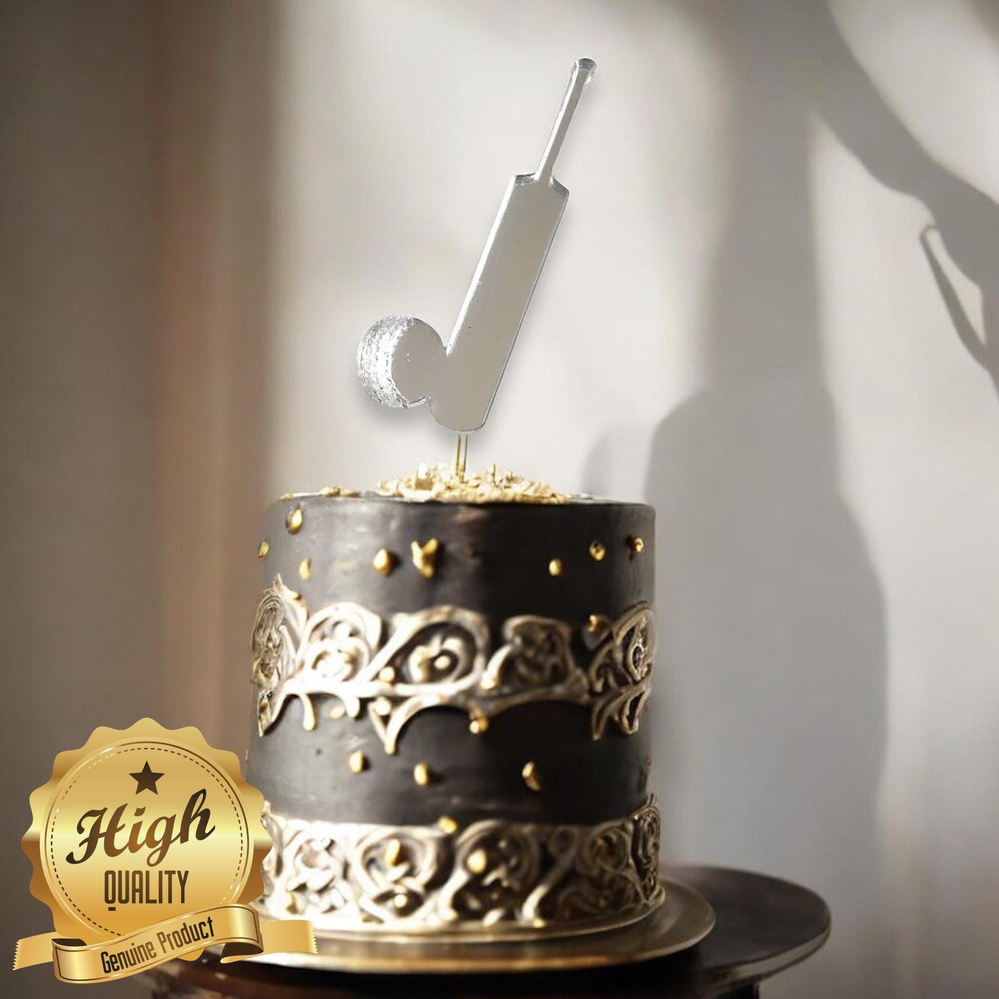A reflective silver cricket bat and ball cake topper atop a black and gold decorative cake.