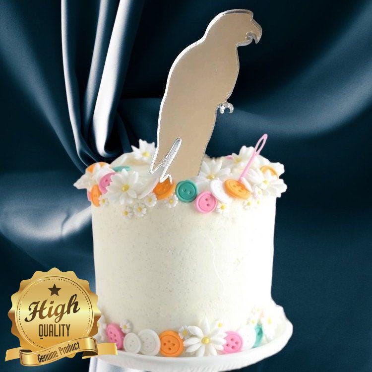 A reflective, mirror-finish parrot silhouette cake topper standing on a white cake decorated with flowers and buttons.