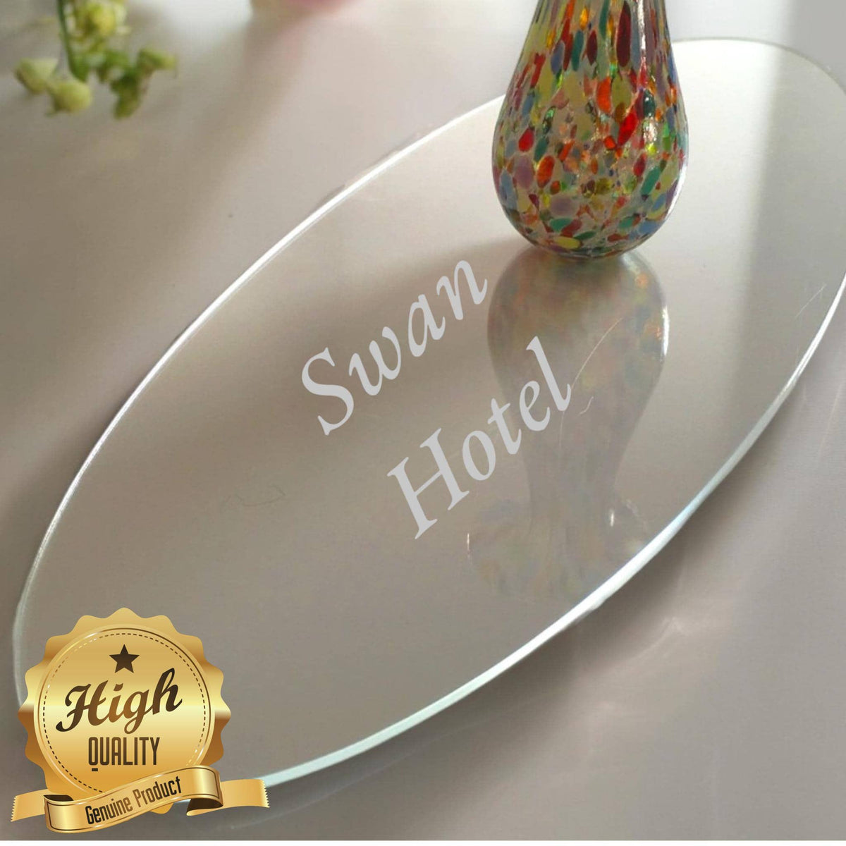 Oval clear acrylic worktop saver engraved with "Swan Hotel," featuring a colorful glass vase on top.
