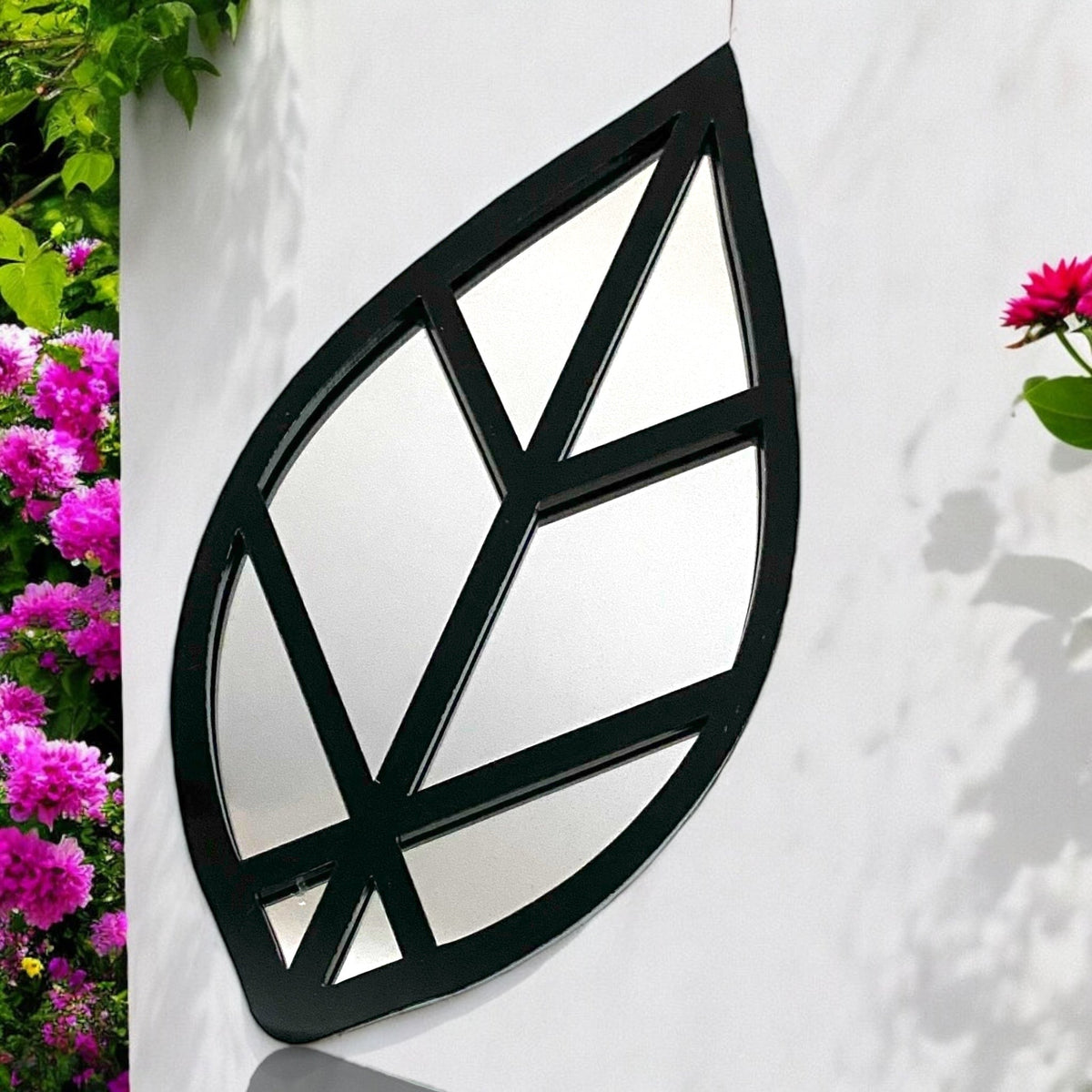 Leaf-shaped garden mirror with a black metal frame, mounted on a white wall.