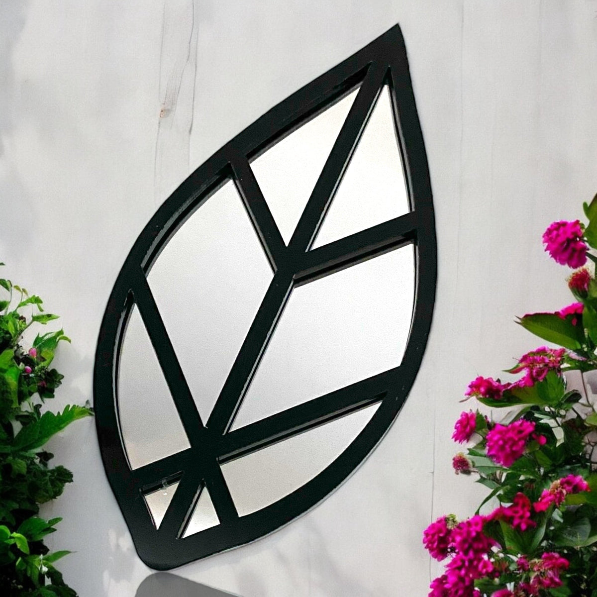 A black leaf-shaped garden mirror hangs on a white wall, flanked by green foliage and pink flowers.