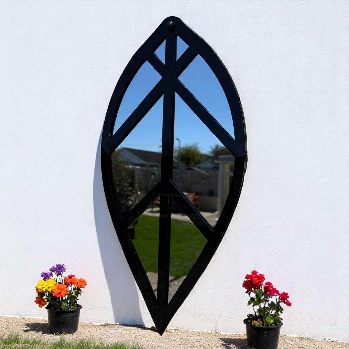Black leaf-shaped garden mirror hanging on a white wall, flanked by potted flowers.