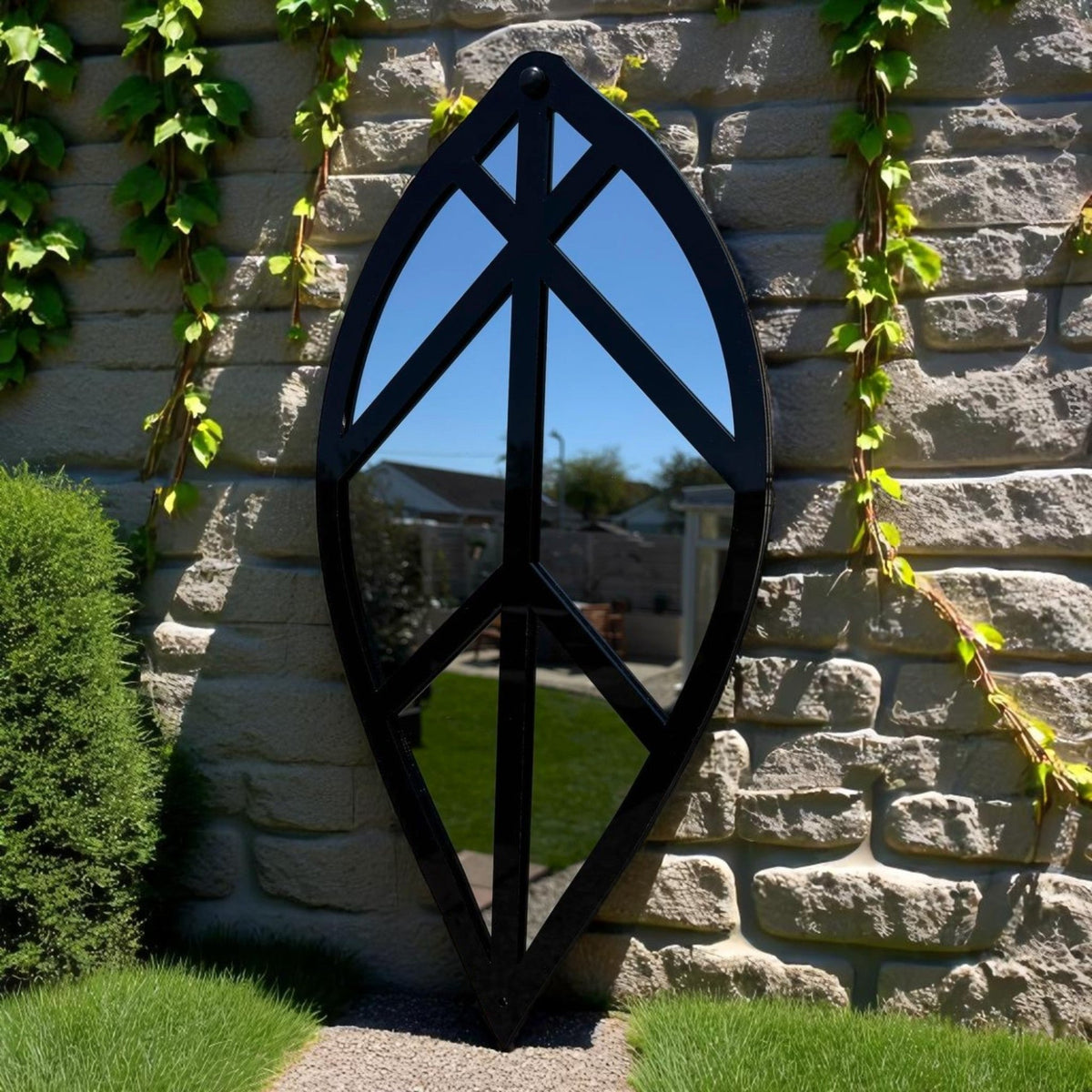 Leaf-shaped garden mirror with a black frame, leaning against a stone wall in a garden.