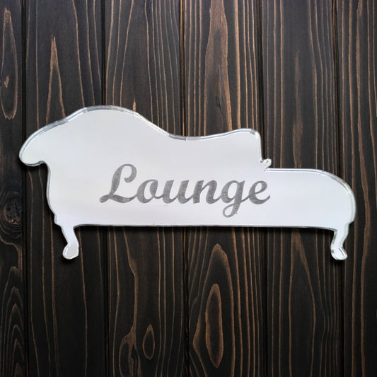 Chai-shaped silver mirror engraved with the word "Lounge" in cursive, on a dark wood background.