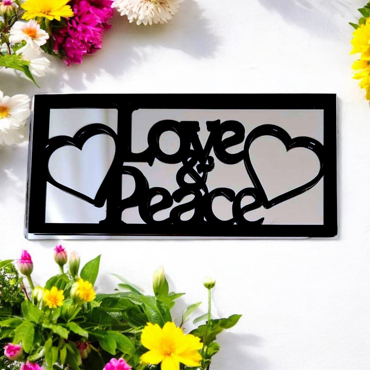 Black framed rectangular garden mirror with "Love & Peace" cut-out text and heart shapes, surrounded by flowers.