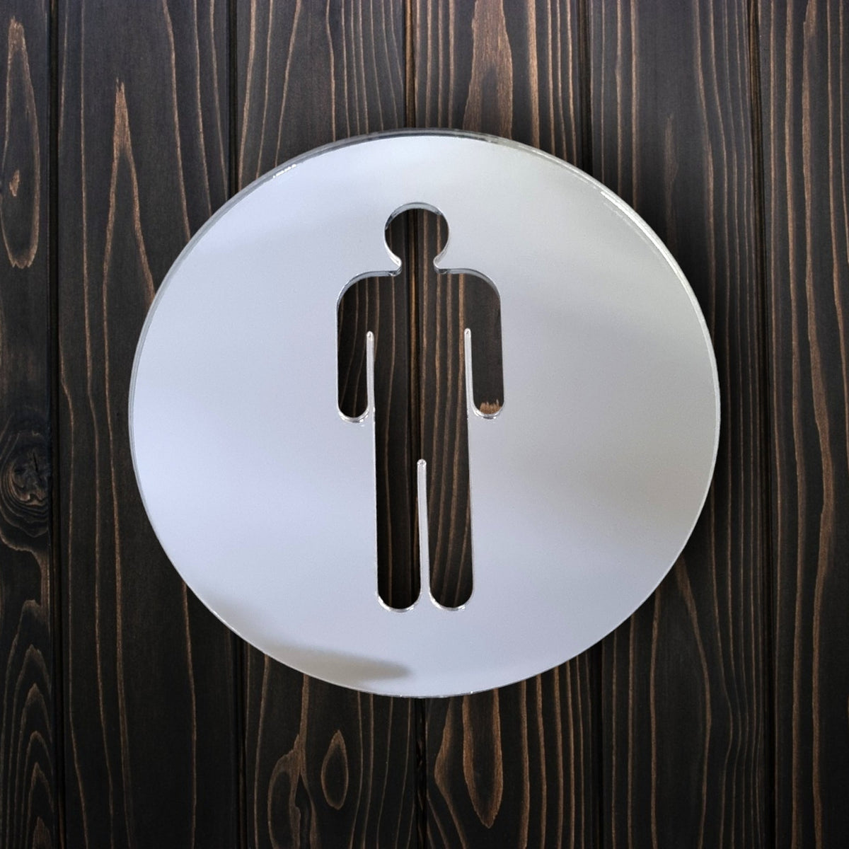 Round mirrored male toilet sign mounted on dark vertical wooden planks.