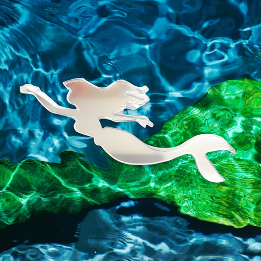 A silver mermaid-shaped acrylic mirror against a blue and green aquatic background.