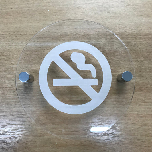 Round clear acrylic no-smoking sign with white engraving and two chrome mounting standoffs on wood.