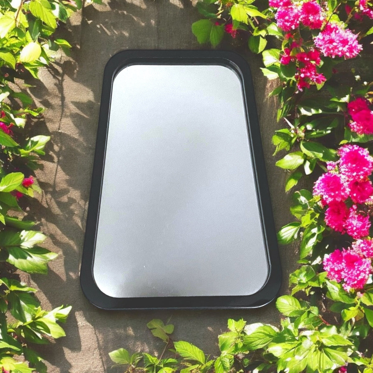 A rectangular garden mirror with a black frame, surrounded by green leaves and bright pink flowers.