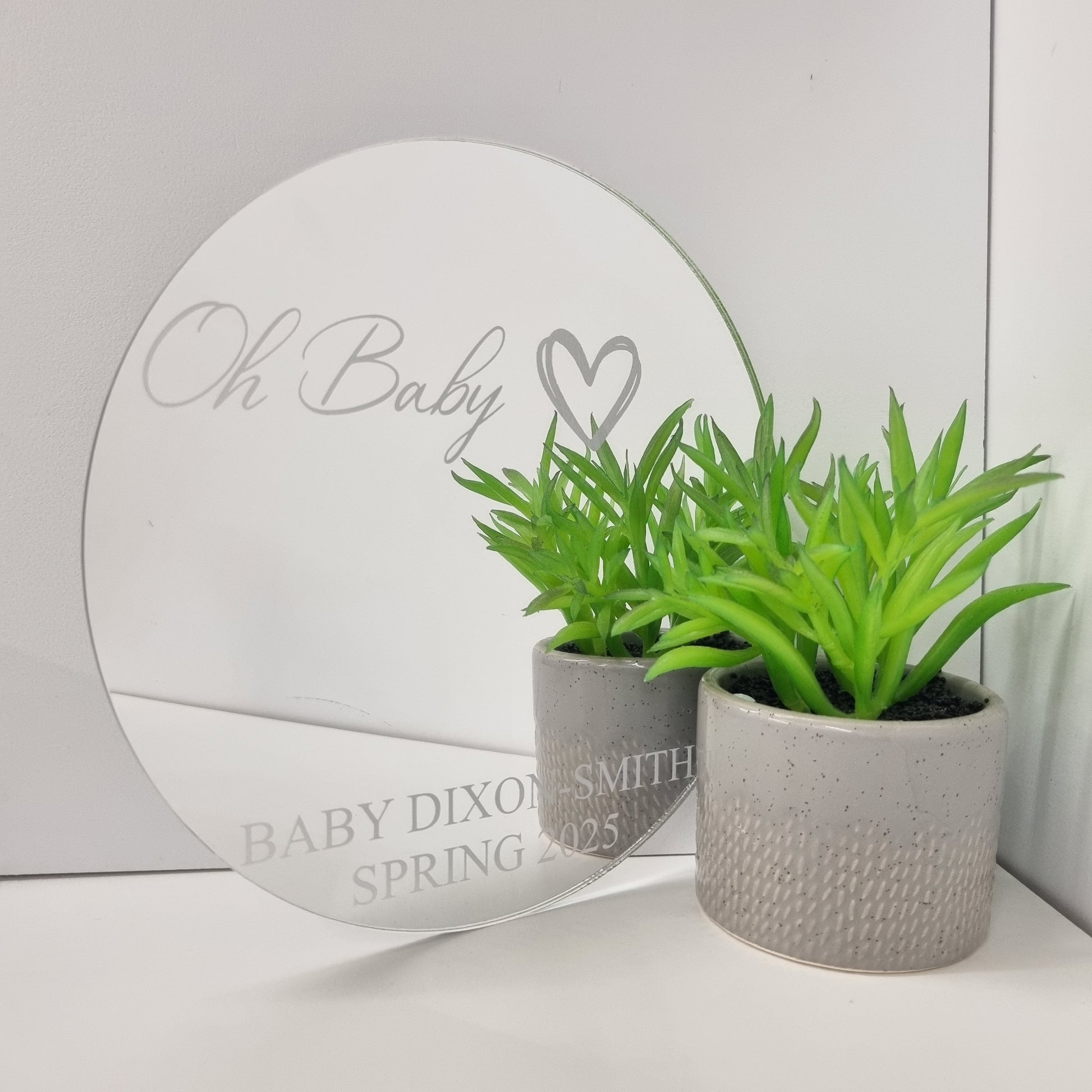 Personalized round mirror with "Oh Baby" and "BABY DIXON-SMITH SPRING 2025" text, next to a potted plant.