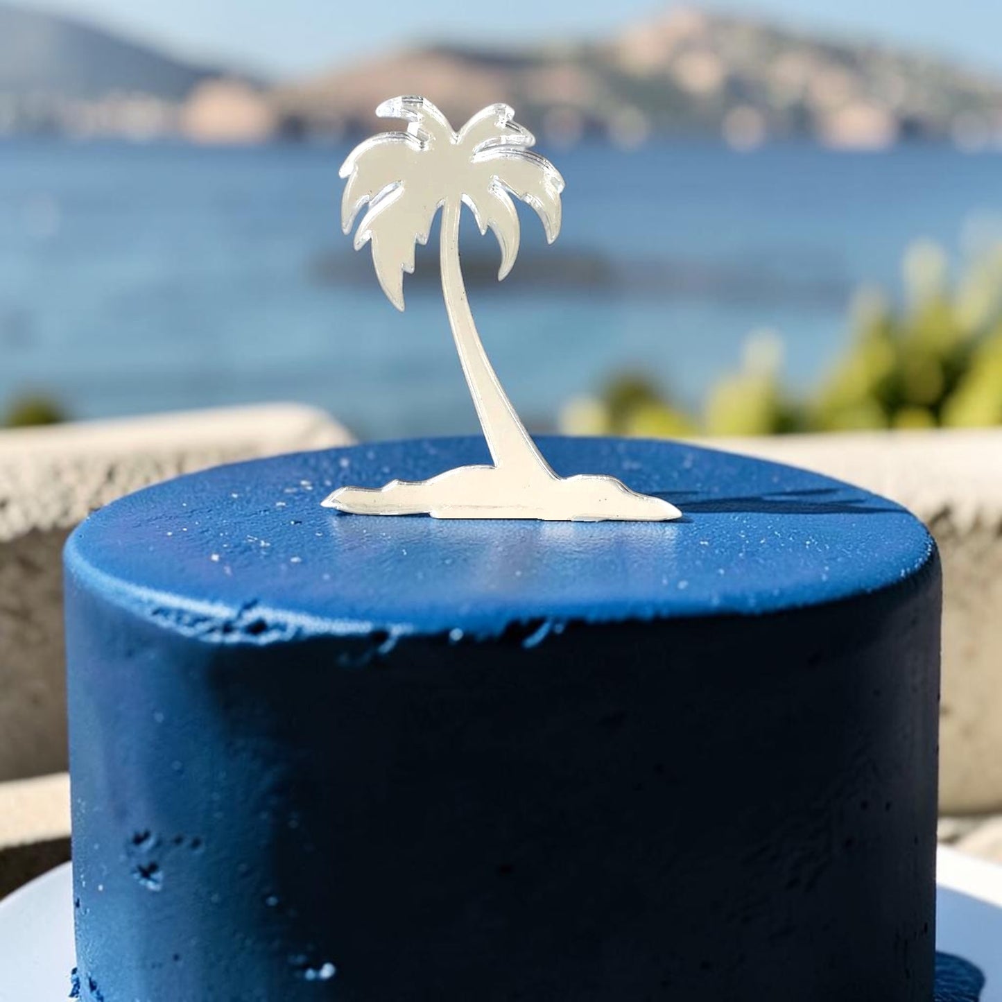 A mirrored, silver palm tree cake topper centered on a round, deep-blue frosted cake against a blurred seaside backdrop.