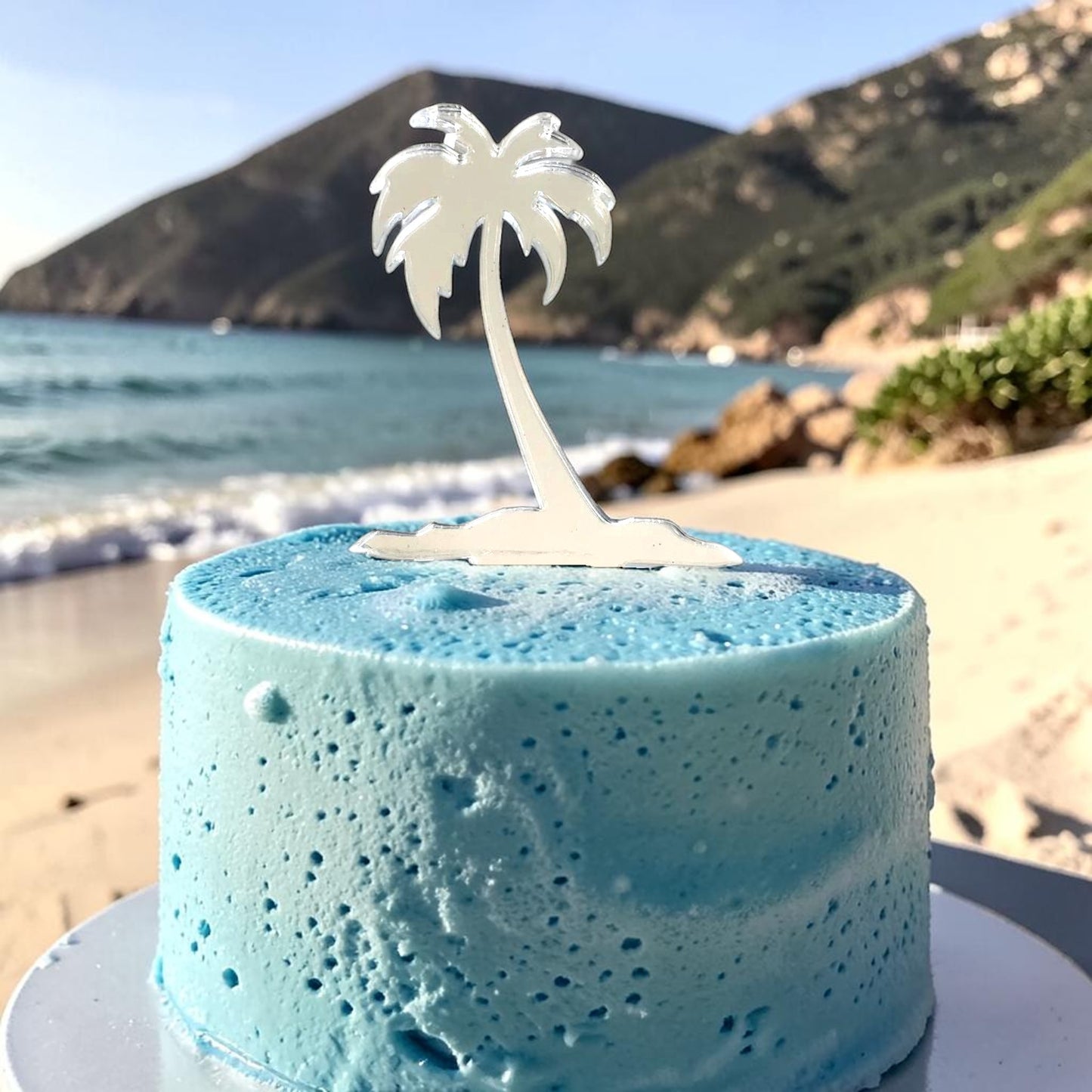 White acrylic palm tree cake topper on a blue, textured cake, set against a beach and mountain background.