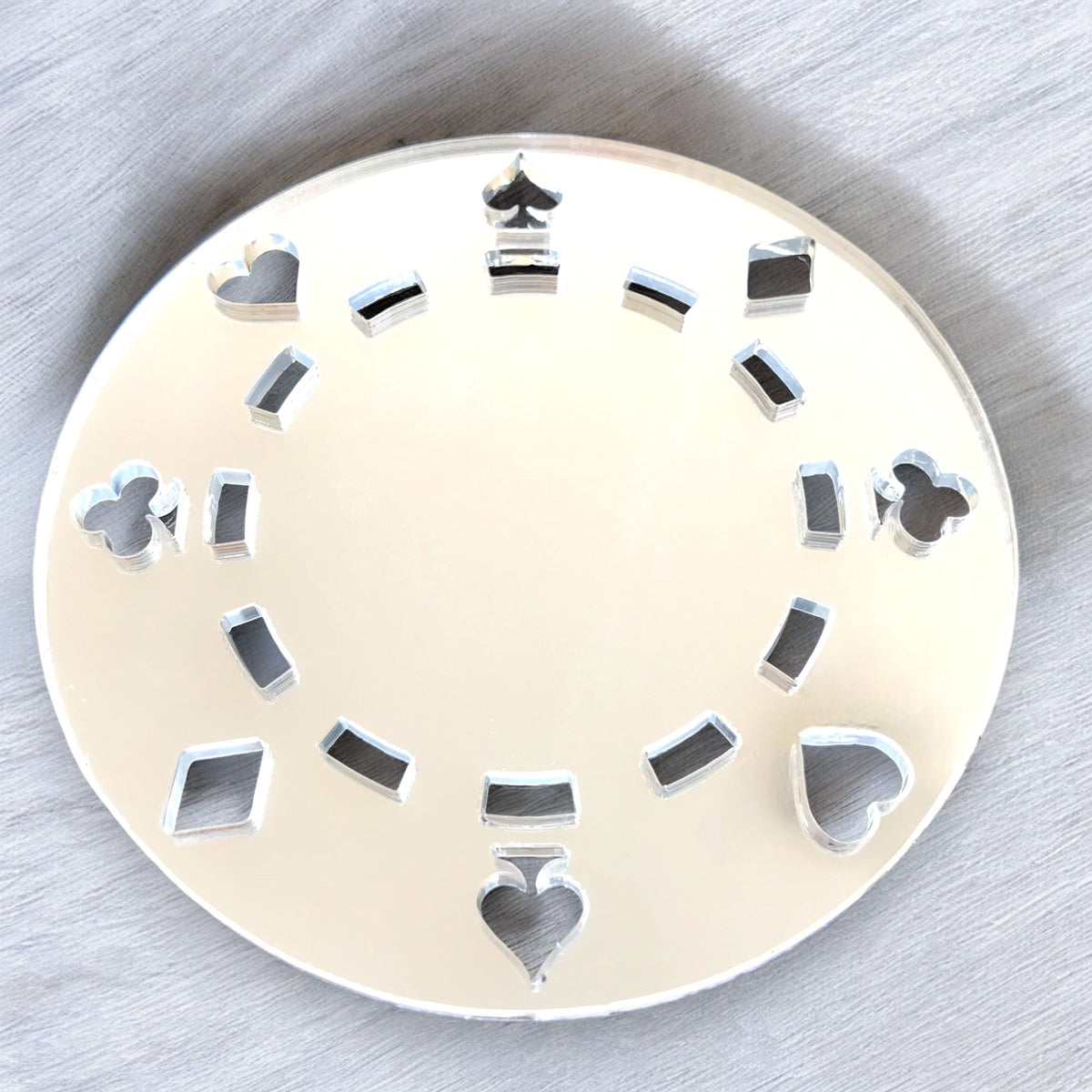 Round silver poker chip-shaped mirror with card suit cutouts along the edge on a grey surface.