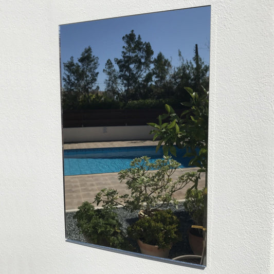 A rectangular garden mirror mounted on a white textured outdoor wall, reflecting a swimming pool and patio plants.