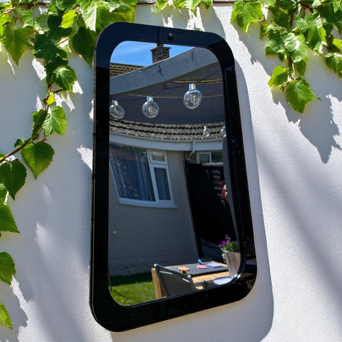 Rectangular black garden mirror mounted on a white outdoor wall with green ivy framing the top and left side.