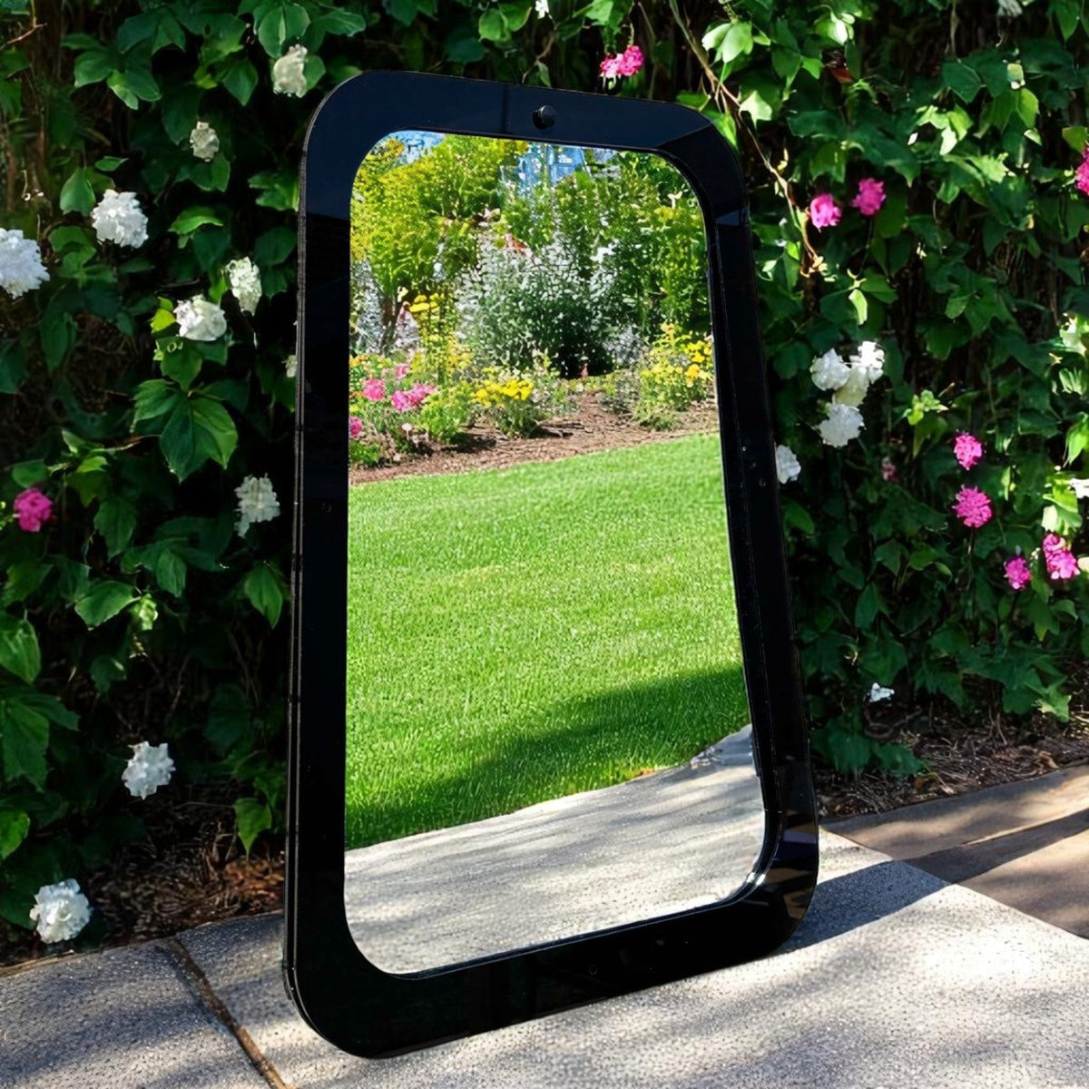 Rectangular garden mirror with a glossy black frame, reflecting a green lawn and flowering bushes outdoors.
