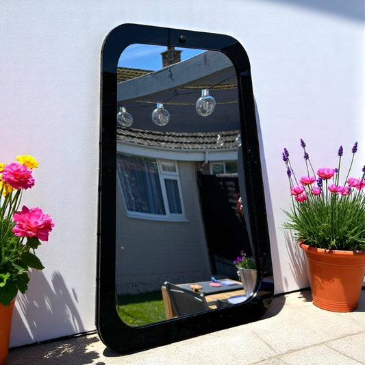 Rectangular garden mirror with a wide black frame, leaning against an outdoor wall between flower pots.