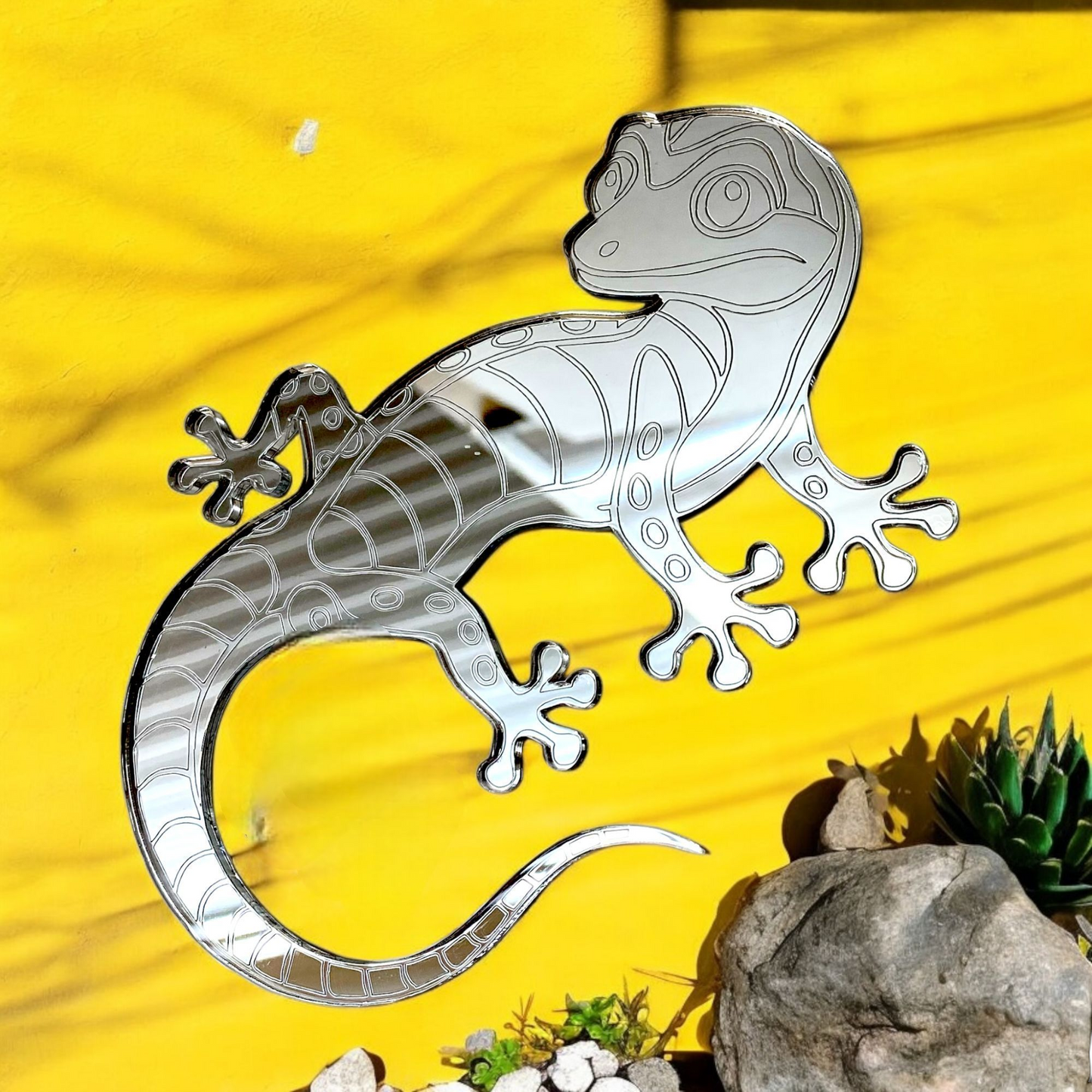 Etched gecko-shaped mirror mounted on a yellow wall, with stones and a small succulent below.