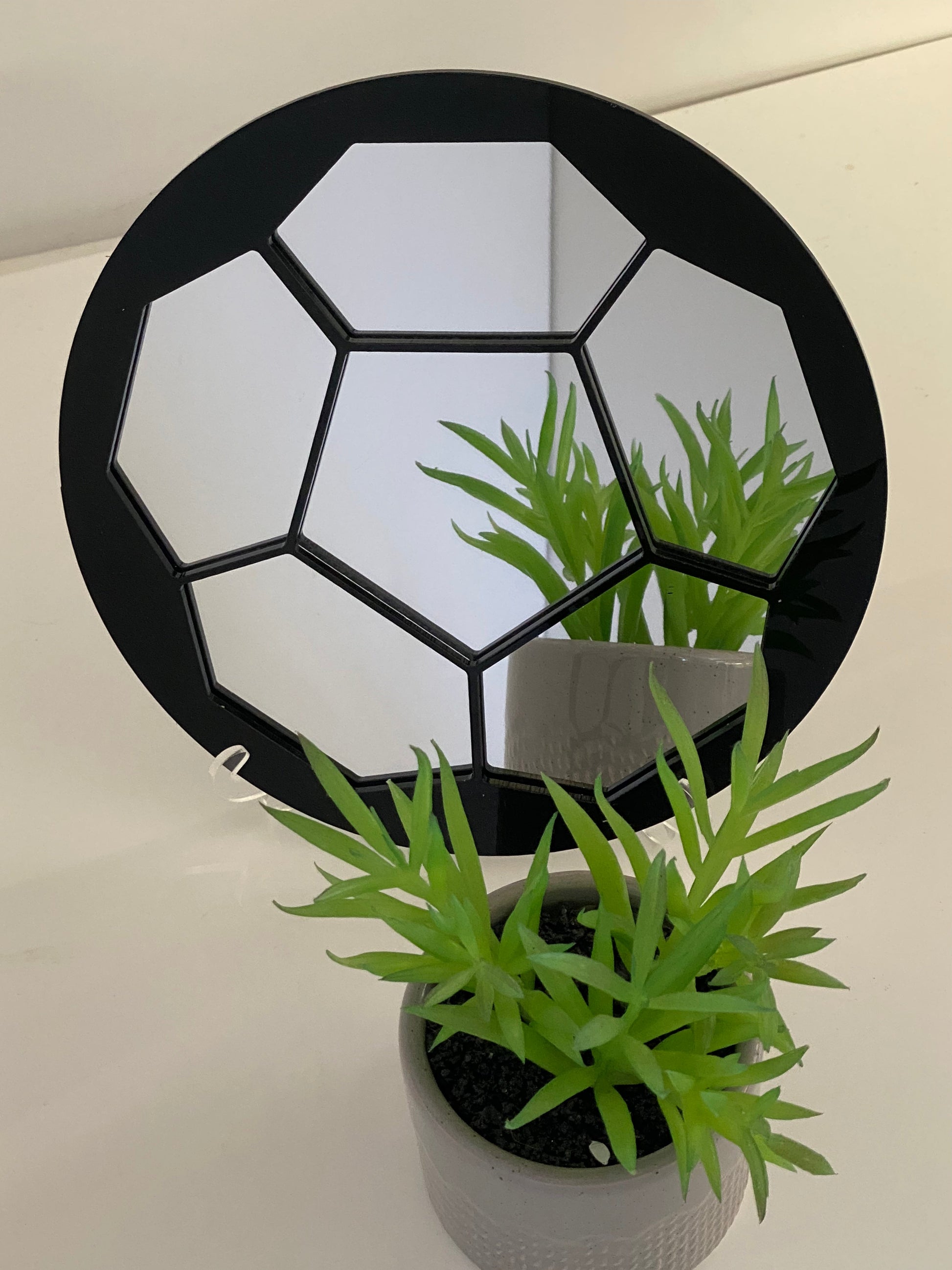 Round black football-patterned framed mirror with segmented panels reflecting a small potted green plant.