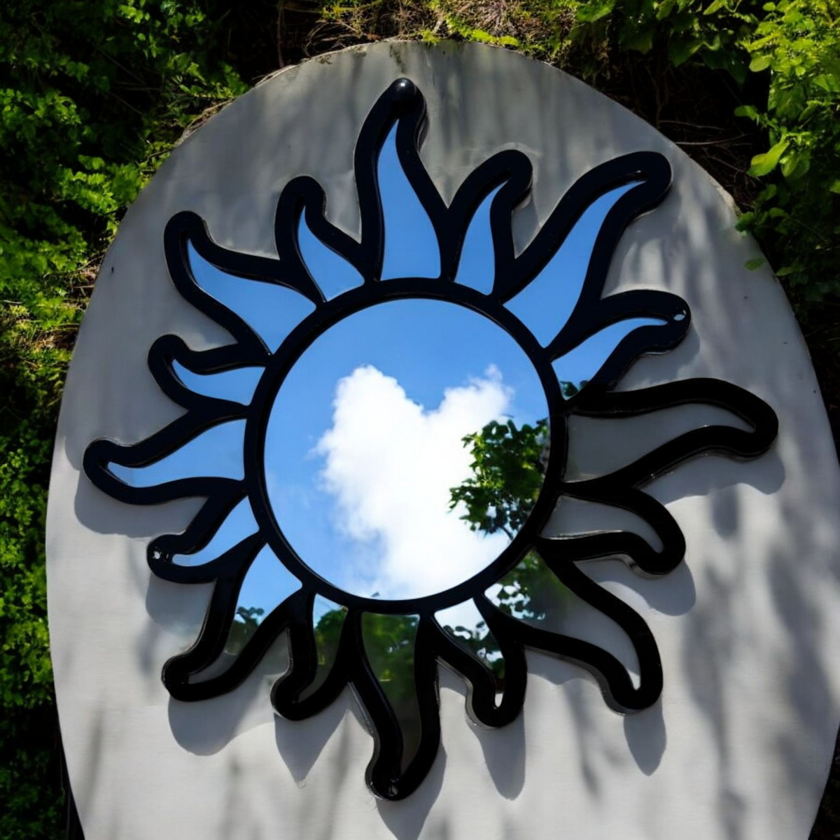 A black, sunburst-framed garden mirror hanging against a light-colored outdoor wall, reflecting the sky and foliage.
