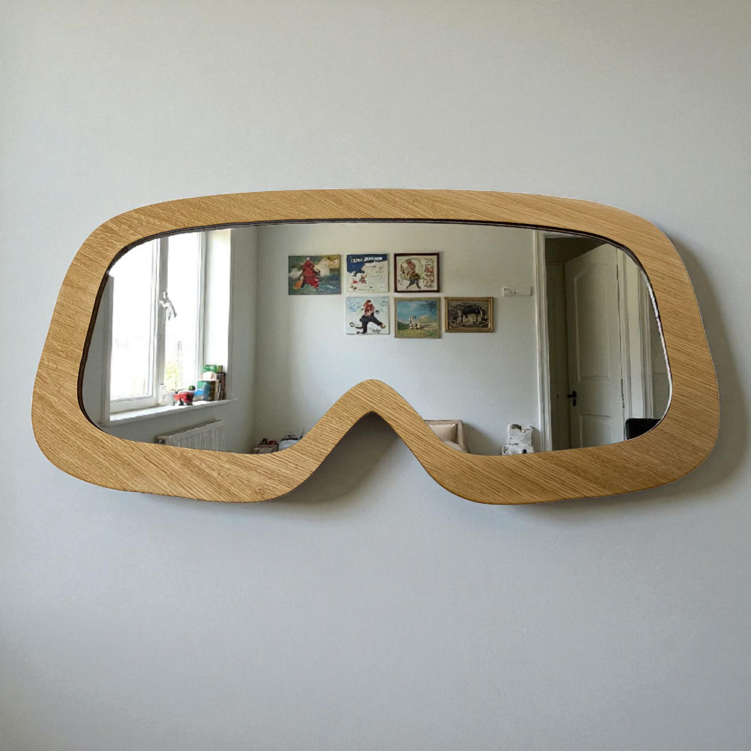 A ski goggle-shaped wall mirror with a light wood grain frame, hanging on a white wall.
