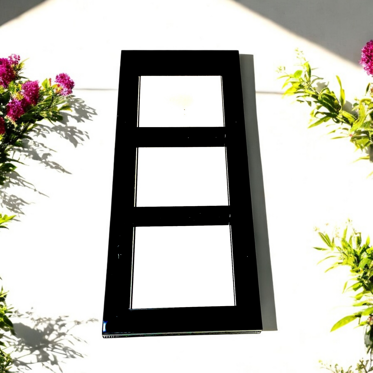 A rectangular three-panel garden mirror with a black frame, flanked by green foliage and purple flowers.