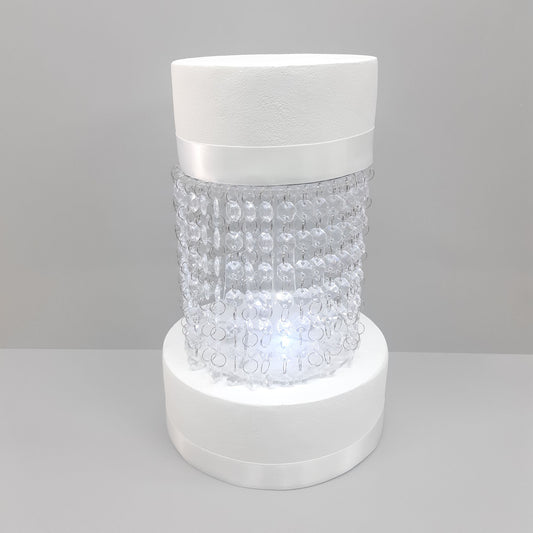 Round acrylic cake separator stand with a central beaded glass section between two white cylindrical bases.
