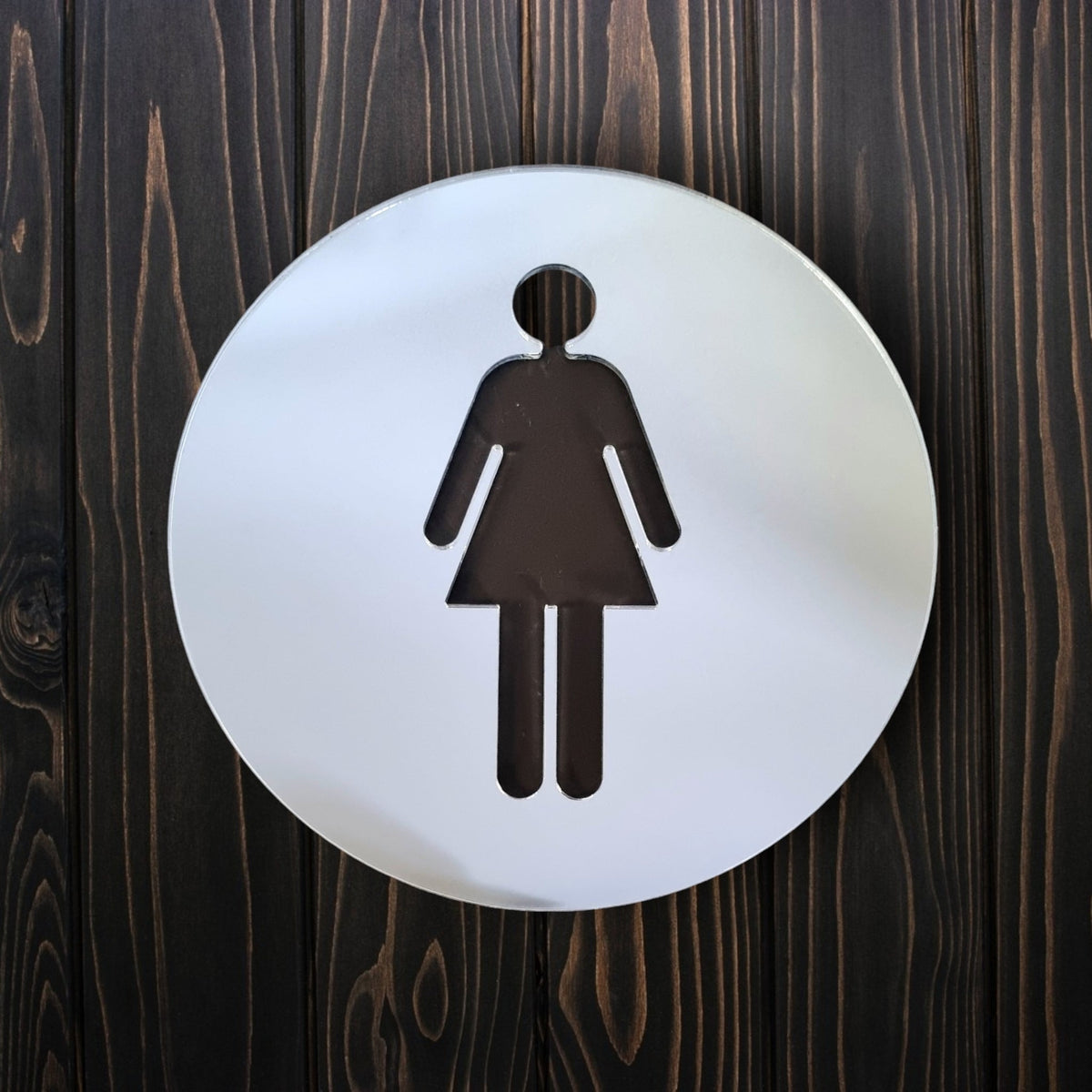 Round mirrored female toilet sign with a cut-out woman icon on a dark wood background.
